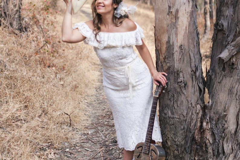 Puede incluir: Una mujer con un vestido blanco de encaje se encuentra en un &aacute;rea boscosa, sosteniendo un sombrero en una mano y una guitarra en la otra. La guitarra es una guitarra resonadora con un agujero de sonido redondo y un cuerpo de metal.