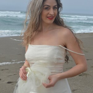 Puede incluir: Una mujer con un vestido blanco sin tirantes con una capa de tul, de pie en una playa de arena con olas del oc&eacute;ano en el fondo.