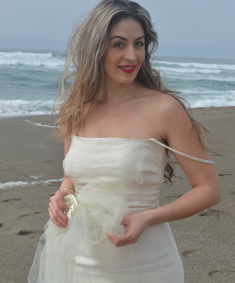 Puede incluir: Una mujer con un vestido blanco sin tirantes con una capa de tul, de pie en una playa de arena con olas del oc&eacute;ano en el fondo.