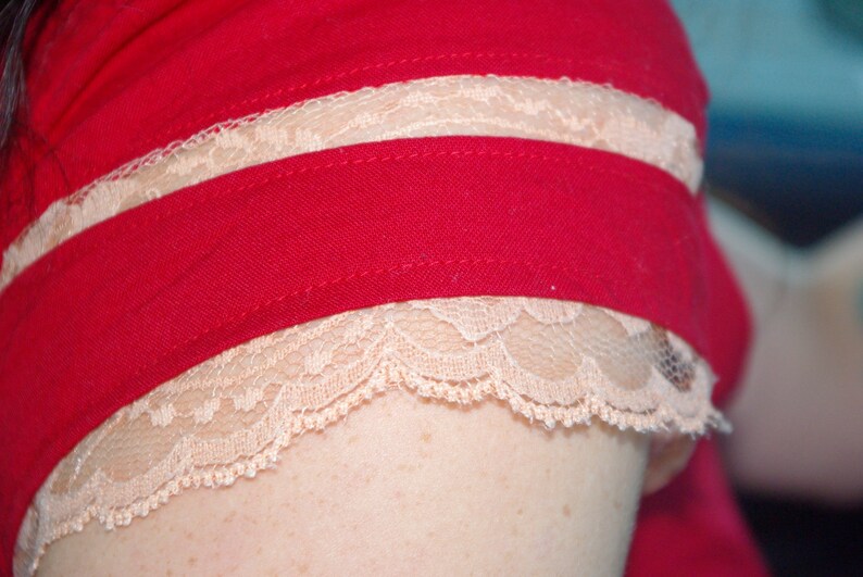 Puede incluir: Una camisa roja con un ribete de encaje blanco alrededor del cuello y las mangas. El ribete de encaje es de doble capa.