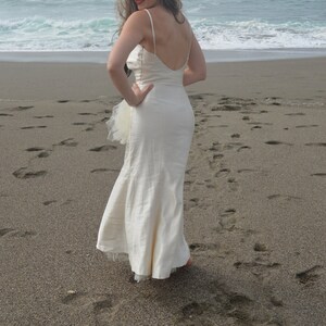 Puede incluir: Una mujer con un vestido blanco sin mangas, largo hasta el suelo, con una capa de tul, est&aacute; de pie en una playa de arena. Est&aacute; de espaldas a la c&aacute;mara y mirando por encima del hombro.