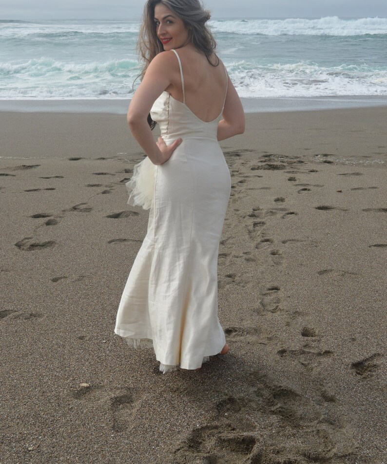 Puede incluir: Una mujer con un vestido blanco sin mangas, largo hasta el suelo, con una capa de tul, est&aacute; de pie en una playa de arena. Est&aacute; de espaldas a la c&aacute;mara y mirando por encima del hombro.