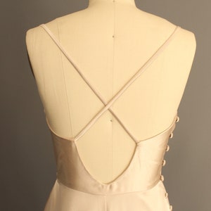 Puede incluir: Un vestido de satén beige claro con la espalda baja y tirantes cruzados. El vestido tiene un cierre de botones en la espalda.