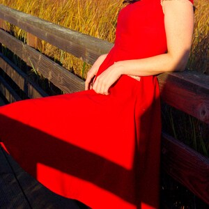 Puede incluir: Una mujer con un vestido rojo con ribetes de encaje blanco se sienta en una barandilla de madera. El vestido tiene un corpi&ntilde;o ajustado y una falda completa. El cabello de la mujer es oscuro y largo. El fondo es un campo de hierba alta.