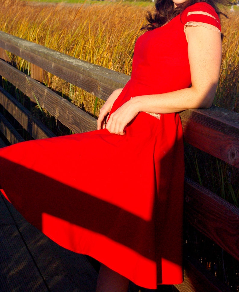 Puede incluir: Una mujer con un vestido rojo con ribetes de encaje blanco se sienta en una barandilla de madera. El vestido tiene un corpi&ntilde;o ajustado y una falda completa. El cabello de la mujer es oscuro y largo. El fondo es un campo de hierba alta.
