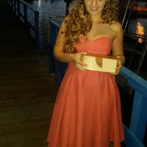 Puede incluir: Una mujer con un sombrero blanco y un vestido de lunares coral sostiene una pequeña caja rectangular de color marrón claro con un cierre dorado.