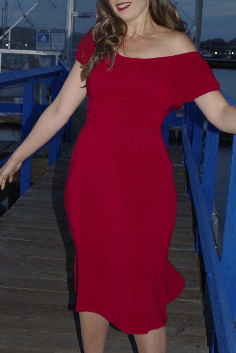 May include: A woman wearing a red off-the-shoulder dress standing on a wooden dock with a blue railing.