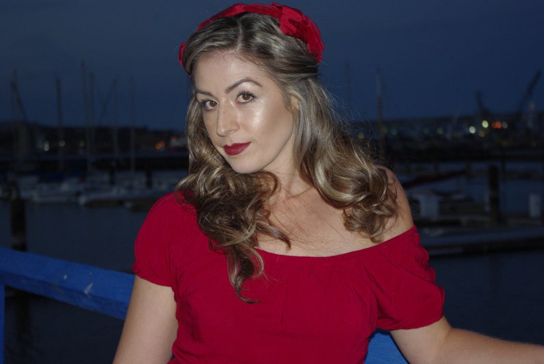 May include: A woman with long blonde hair wearing a red off-the-shoulder dress and a red headband. She is standing in front of a blue background.