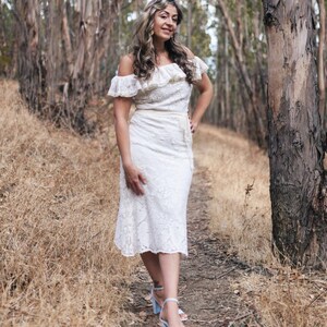 Puede incluir: Una mujer con un vestido blanco de encaje de hombros descubiertos con un escote con volantes y un cintur&oacute;n. Est&aacute; de pie en un entorno de bosque con &aacute;rboles y hierba seca.