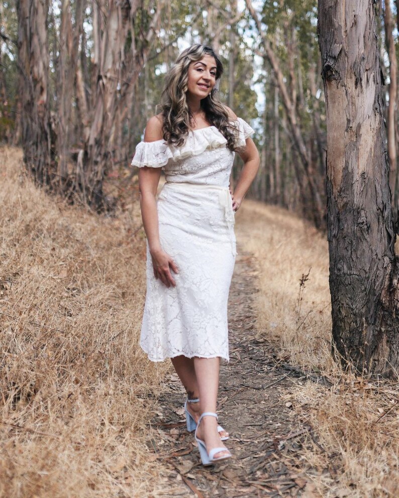 Puede incluir: Una mujer con un vestido blanco de encaje de hombros descubiertos con un escote con volantes y un cintur&oacute;n. Est&aacute; de pie en un entorno de bosque con &aacute;rboles y hierba seca.