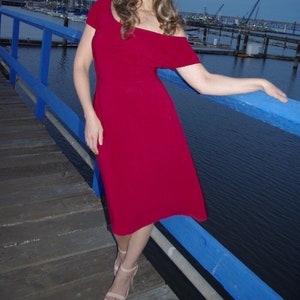 May include: A woman wearing a bright red off-the-shoulder midi dress with short sleeves, standing on a wooden dock with a blue railing. She is wearing nude heels and has long brown hair.