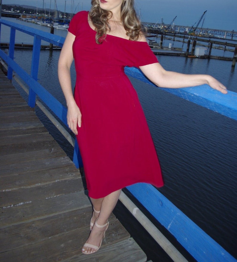 May include: A woman wearing a bright red off-the-shoulder midi dress with short sleeves, standing on a wooden dock with a blue railing. She is wearing nude heels and has long brown hair.