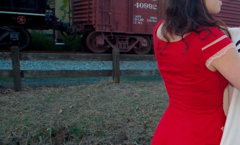 Puede incluir: Una mujer con un vestido rojo con un ribete de encaje rosa en las mangas. El vestido tiene un escote redondo y es sin mangas. La mujer est&aacute; de pie frente a un vag&oacute;n de tren rojo con el n&uacute;mero 40992 en el lateral.
