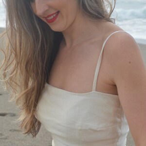 Puede incluir: Una mujer con el pelo largo y ondulado casta&ntilde;o lleva un top blanco sin mangas de cuello cuadrado. Est&aacute; de pie en una playa de arena con el oc&eacute;ano de fondo.