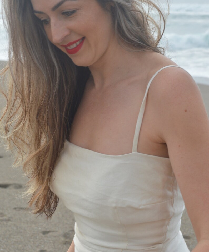 Puede incluir: Una mujer con el pelo largo y ondulado casta&ntilde;o lleva un top blanco sin mangas de cuello cuadrado. Est&aacute; de pie en una playa de arena con el oc&eacute;ano de fondo.