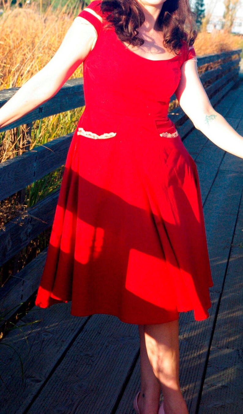 Puede incluir: Una mujer con un vestido rojo con ribetes de encaje blanco y un corpi&ntilde;o ajustado. El vestido tiene una falda completa y ondea con el viento. Est&aacute; de pie en una pasarela de madera.