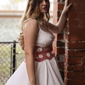 Puede incluir: Una mujer con un vestido blanco y un cinturón floral rosa se encuentra junto a una pared de ladrillo. Está mirando hacia un lado y tiene la mano en la pared.