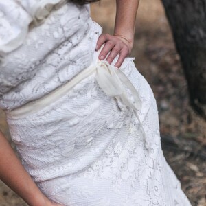 Puede incluir: Un vestido blanco de encaje con un delicado patr&oacute;n floral y una cinta blanca atada a la cintura.
