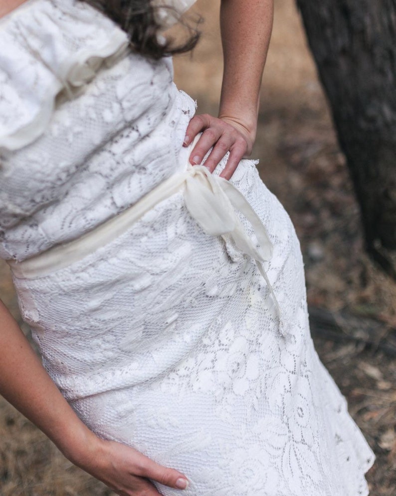 Puede incluir: Un vestido blanco de encaje con un delicado patr&oacute;n floral y una cinta blanca atada a la cintura.