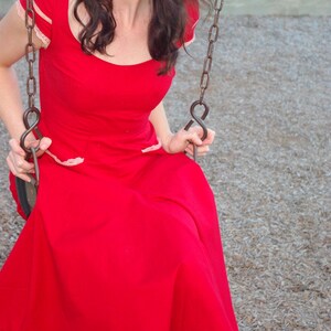 Puede incluir: Una mujer con un vestido rojo con ribete de encaje blanco se sienta en un columpio. El columpio est&aacute; hecho de cadenas met&aacute;licas y tiene un asiento de madera.