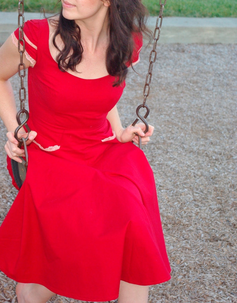 Puede incluir: Una mujer con un vestido rojo con ribete de encaje blanco se sienta en un columpio. El columpio est&aacute; hecho de cadenas met&aacute;licas y tiene un asiento de madera.