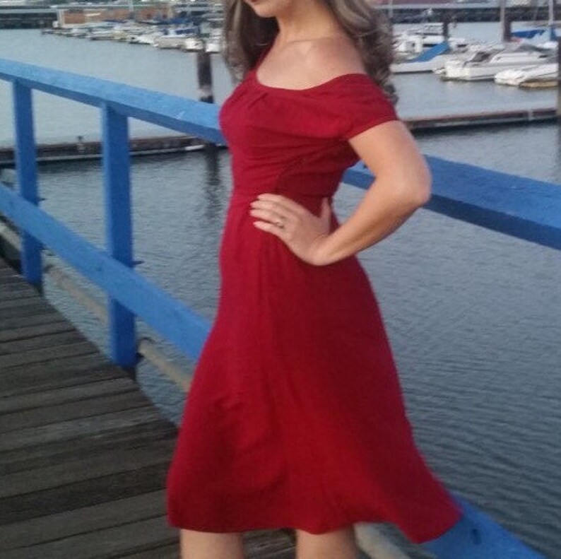 May include: A woman wearing a red off-the-shoulder dress with short sleeves. She is standing on a wooden dock with a blue railing.