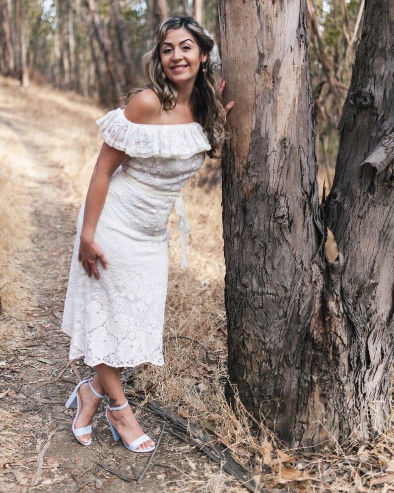 Puede incluir: Una mujer con un vestido blanco de encaje con un escote de volantes se encuentra en un entorno boscoso. Lleva sandalias de tac&oacute;n azul claro y tiene el pelo recogido hacia atr&aacute;s.