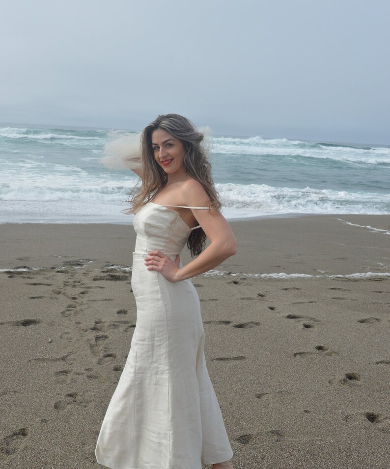 Puede incluir: Una mujer con un vestido de lino blanco se encuentra en una playa de arena con el oc&eacute;ano de fondo. El vestido tiene un dise&ntilde;o simple y elegante con tirantes finos y una falda fluida.