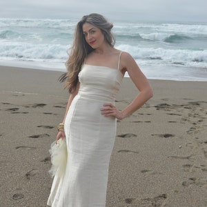 Puede incluir: Una mujer con un vestido blanco de lino est&aacute; de pie en una playa de arena con olas del oc&eacute;ano en el fondo.