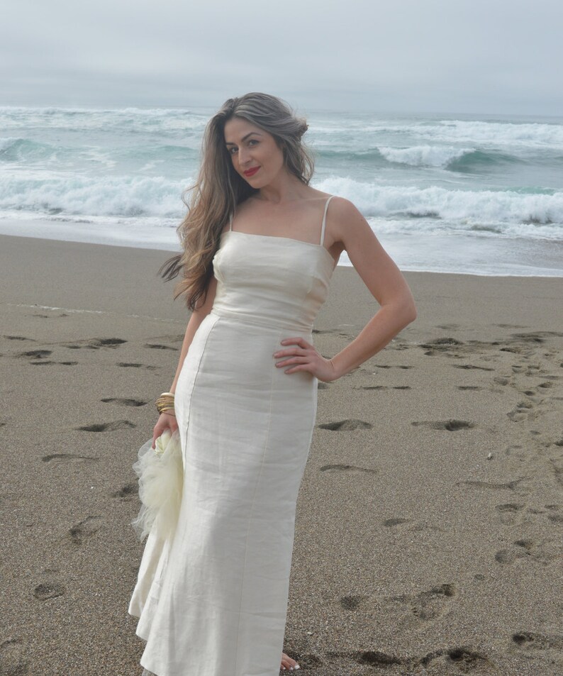 Puede incluir: Una mujer con un vestido blanco de lino est&aacute; de pie en una playa de arena con olas del oc&eacute;ano en el fondo.