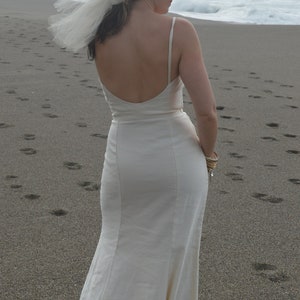 Puede incluir: Una mujer con un vestido de novia blanco sin mangas y falda fluida. Est&aacute; de pie en una playa de arena con el oc&eacute;ano de fondo.