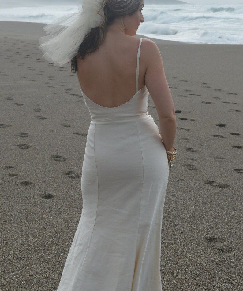 Puede incluir: Una mujer con un vestido de novia blanco sin mangas y falda fluida. Est&aacute; de pie en una playa de arena con el oc&eacute;ano de fondo.