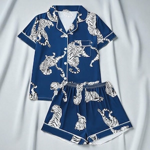 Puede incluir: Conjunto de pijama de dos piezas en azul marino con estampado de tigre blanco. La parte superior de manga corta y los pantalones cortos tienen ribetes blancos en los bordes. La parte superior tiene botones y un bolsillo en el pecho.