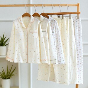 May include: Cream-colored pajama sets hanging on a wooden rack. The long-sleeved shirts and pants feature a small floral pattern and are trimmed with pink or green piping. The fabric appears soft and lightweight, suitable for sleepwear.