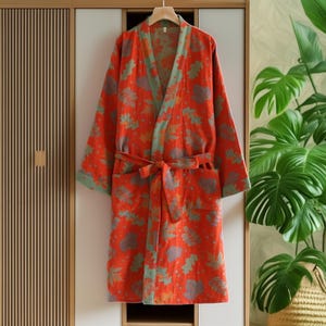 May include: An orange robe with a floral pattern in shades of green, grey, and yellow. The robe has a matching belt, pockets, and green trim along the collar and cuffs. The robe is hanging on a wooden hanger.