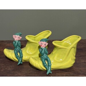 May include: A pair of ceramic elf boot planters in a bright yellow-green color. Each boot has a seated elf figurine with green clothing and pink skin. The planters are likely vintage and suitable for small plants.