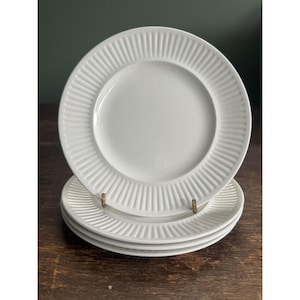 May include: A stack of four white ceramic plates. Each plate has a ridged, textured rim and a smooth, circular center. The plates are stacked on a wooden surface, with a dark green background.