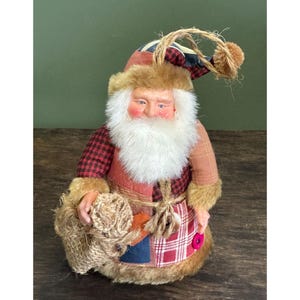 May include: A fabric Santa Claus figurine with a white beard, rosy cheeks, and a plaid coat. The Santa Claus is holding a burlap sack and wearing a hat with a loop for hanging. The figurine has a rustic, handmade appearance.