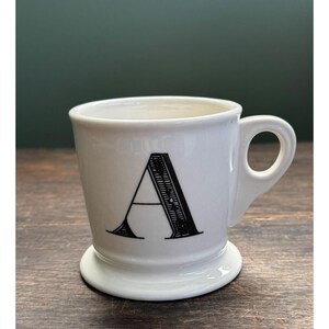 K&ouml;nnte beinhalten: Wei&szlig;er Keramikbecher mit dem schwarzen Buchstaben "A" auf der Vorderseite. Der Becher hat einen abgerundeten Boden und einen Henkel. Der Becher steht auf einer Holzoberfl&auml;che.