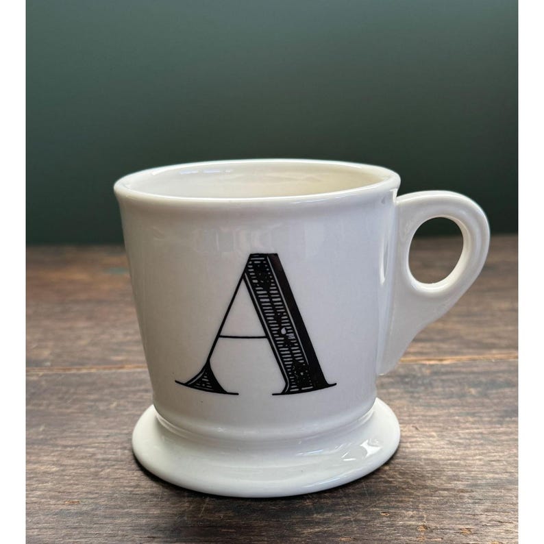 K&ouml;nnte beinhalten: Wei&szlig;er Keramikbecher mit dem schwarzen Buchstaben "A" auf der Vorderseite. Der Becher hat einen abgerundeten Boden und einen Henkel. Der Becher steht auf einer Holzoberfl&auml;che.