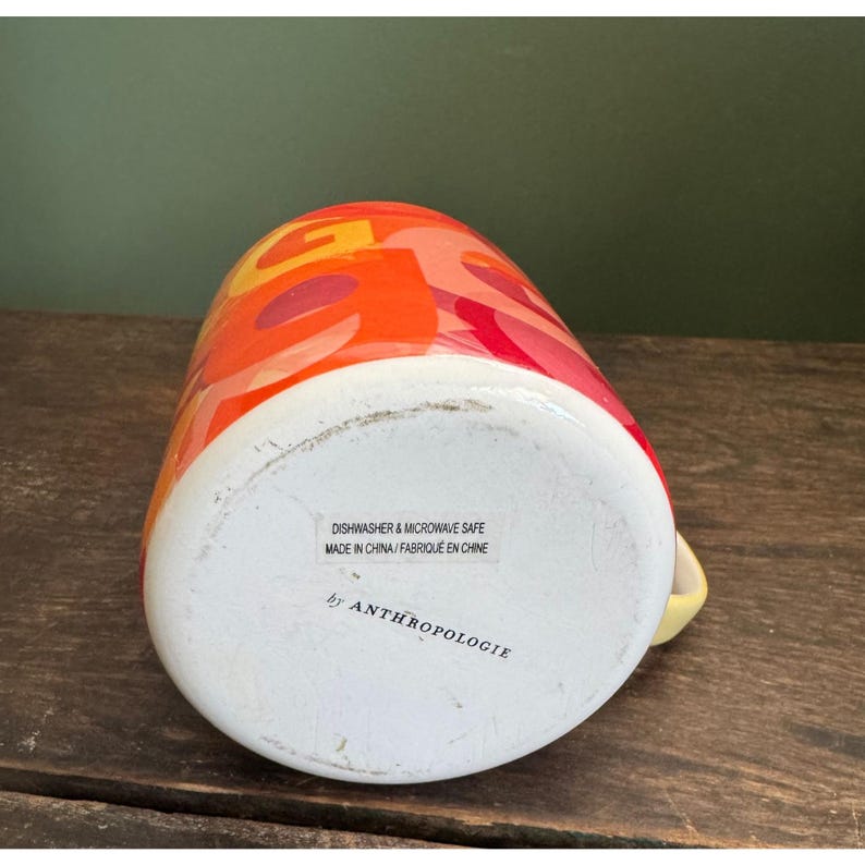 May include: A ceramic mug with a colorful abstract design in shades of orange, red, and yellow. The mug has a white base and a yellow handle. The text "DISHWASHER & MICROWAVE SAFE" and "by ANTHROPOLOGIE" are visible.