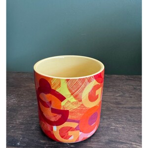 May include: A cylindrical ceramic planter with a yellow interior and a vibrant exterior design. The exterior features a repeating pattern of the letter "G" in shades of red, orange, and yellow, creating a bold, artistic look.