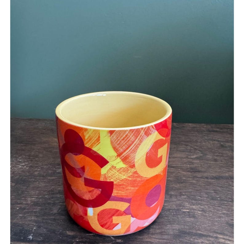 May include: A cylindrical ceramic planter with a yellow interior and a vibrant exterior design. The exterior features a repeating pattern of the letter "G" in shades of red, orange, and yellow, creating a bold, artistic look.