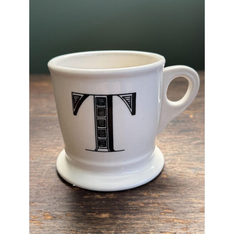 Op de afbeelding: Een witte keramische mok met een zwart handvat en de tekst "But First, Coffee" in een zwarte, vette letter. De mok staat op een wit oppervlak. De mok is ongeveer 10 cm hoog.