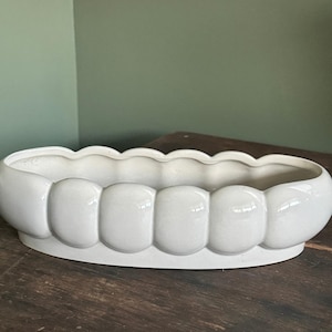 May include: An elongated, oval-shaped ceramic planter with a glossy white finish. The exterior features a repeating pattern of rounded, bulbous segments along the sides, creating a scalloped top edge and a smooth, continuous base structure.