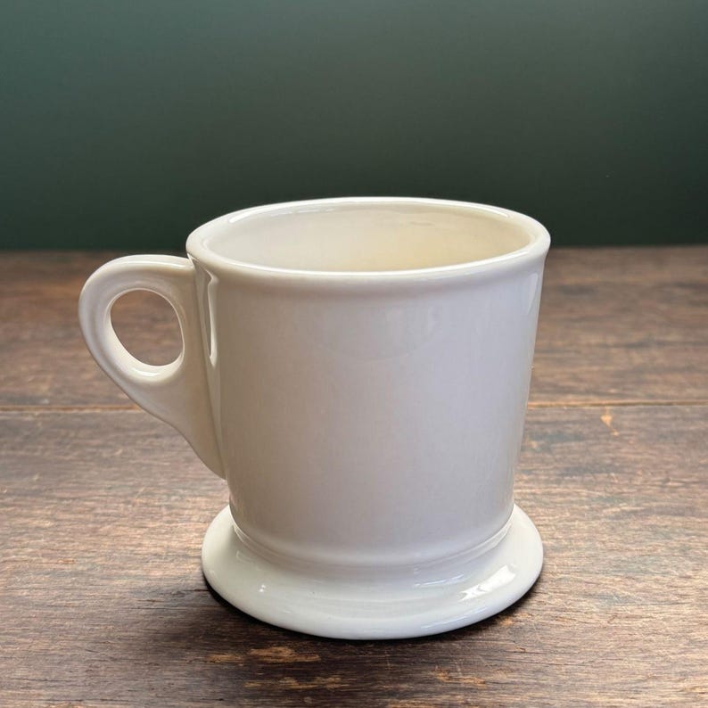 K&ouml;nnte beinhalten: Eine wei&szlig;e Keramik-Tasse mit rundem Boden und Henkel. Die Tasse hat ein schlichtes, klassisches Design und eignet sich f&uuml;r Kaffee, Tee oder andere Getr&auml;nke. Die Tasse steht auf einer Holzoberfl&auml;che.
