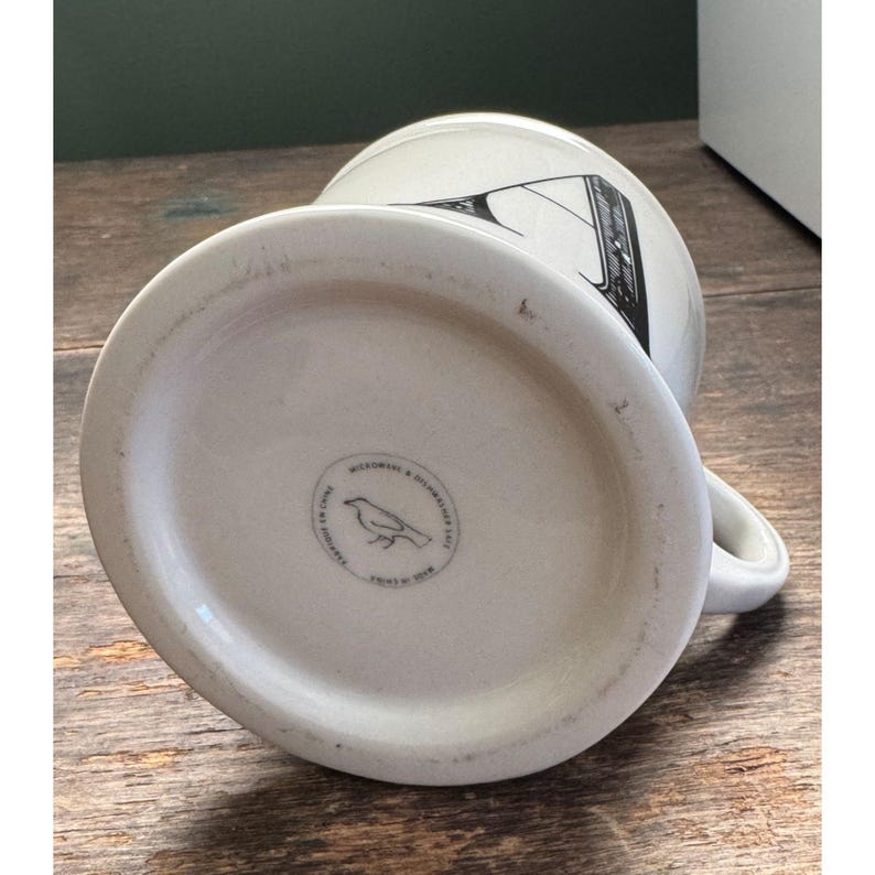 K&ouml;nnte beinhalten: Cremefarbene Keramiktasse mit schwarzem Grafikdesign. Der Boden der Tasse hat einen runden Stempel mit einer Vogelillustration und Text. Die Tasse hat einen Henkel und ist f&uuml;r Hei&szlig;getr&auml;nke konzipiert.