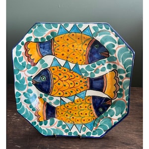 Puede incluir: Una fuente de cerámica pintada a mano con forma octogonal. La fuente presenta tres peces estilizados en amarillo, naranja y azul, sobre un fondo de follaje verde y blanco. Los peces tienen aletas azules y están decorados con puntos naranjas.
