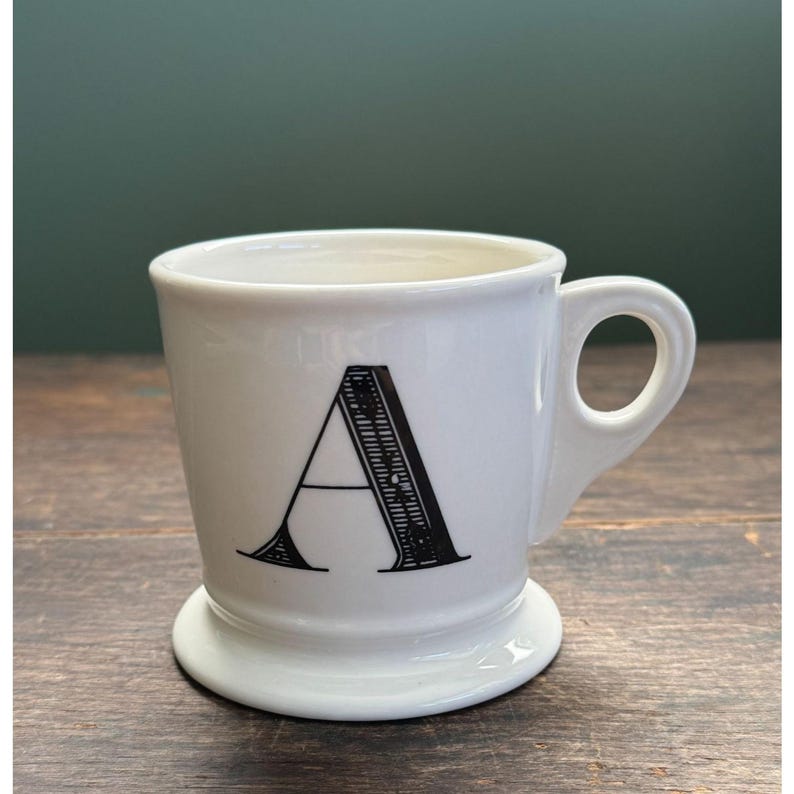 K&ouml;nnte beinhalten: Wei&szlig;e Keramiktasse mit dem schwarzen Buchstaben "A" auf der Vorderseite. Die Tasse hat einen abgerundeten Boden und einen Henkel. Der Hintergrund ist eine dunkelgr&uuml;ne Wand und ein Holztisch.