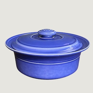May include: A round, cobalt blue ceramic casserole dish with a matching lid. The dish features a smooth, glossy finish, a wide rim, and a central knob on the lid. The design includes concentric circular ridges on the lid and the body of the vessel.
